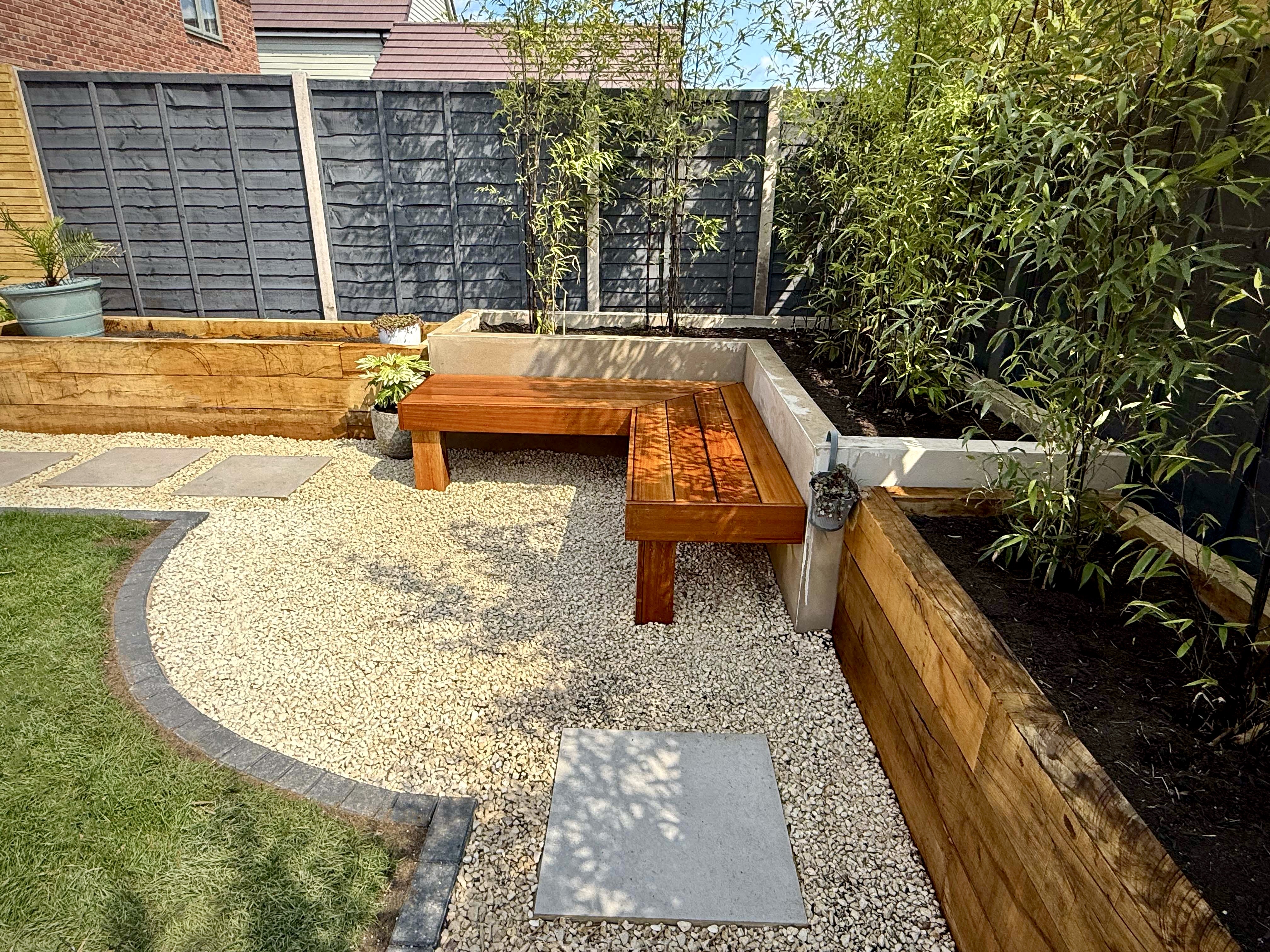 A curved gravel seating area with a bespoke hardwood bench, raised timber planters and evergreen screening planting in a Sussex garden by Brunswick Landscapes